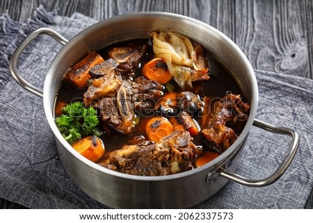 beef broth of beef meat on bones slow cooked with charred vegetables: carrot, onion, garlic, and spices served in a pot on a wooden table, top view
