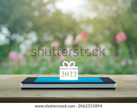 Gift box happy new year 2022 flat icon on modern smart mobile phone screen on wooden table over blur pink flower and tree in park, Business happy new year 2022 shop online concept