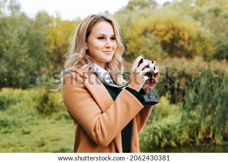 Charming young woman photographer with vintage camera standing in park outdoors. Smiling hipster girl in coat practicing hobby retro film photo.