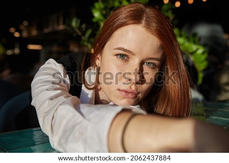 young caucasian stylish redhead pretty woman with long hair lays her head on her arms and table. beautiful pensive melancholic red headed lady laying at sunny cafe. lifestyle portrait