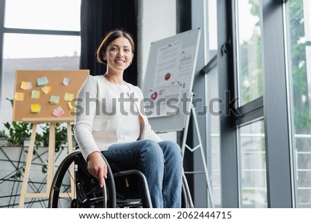cheerful businesswoman in wheelchair looking at camera near blurred infographics in office Royalty-Free Stock Photo #2062444715