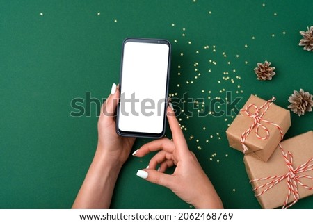 Female hands holding modern smartphone with mockup on green background with christmas decoration. Flat lay template. Fir wreath, gift box