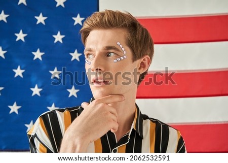 Queer man with rhinestones near his eyes touching face with fingers and looking dreamy