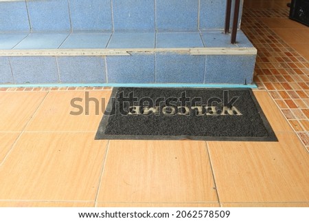 A black doormat sits on the steps of the stairs which are lined with brown tiles. The stairs are paved with blue tiles. There is a white letter "Welcome" on it.