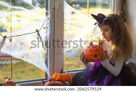 A girl in a witch costume on the windowsill by the window at a Halloween party, decorating the house cobwebs, bats, spiders, pumpkins. Terrible scenery, fear and horror