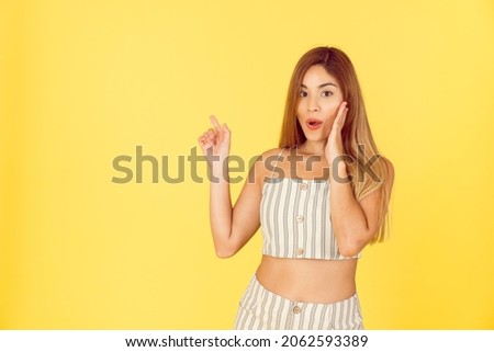 Young latin woman pointing to text, smile and surprise on yellow background. 