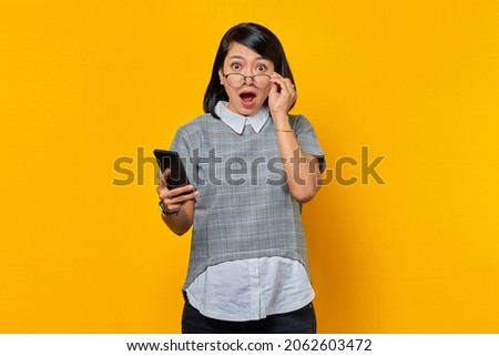 Portrait of Shocked and Suprised young Asian woman holding smartphone and eyewear looking at camera over yellow background Royalty-Free Stock Photo #2062603472