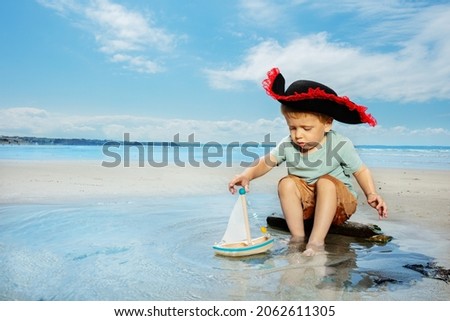 Little pirate blond cute boy play with a boat in the puddle on the sand sea beach and smiling