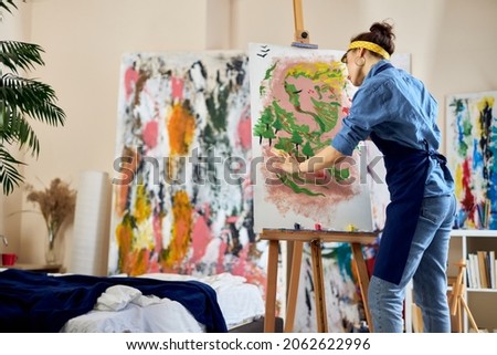 Creative woman, female artist in apron looking inspired while creating her own abstract painting at home studio workshop Royalty-Free Stock Photo #2062622996