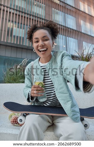 Youth generation and hobby concept. Positive young female model makes selfie after riding skateboard drinks water feels thirsty expresses happy emotions poses against urban setting near publicity area Royalty-Free Stock Photo #2062689500