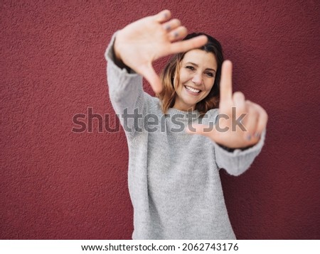 Friendly creative young woman making a frame gesture with her hands framing her face as she visualises a new idea or project standing against a red wall with copyspace and vignette Royalty-Free Stock Photo #2062743176