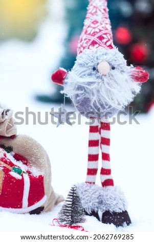 Snowy handmade dwarf close up in red white clothes with bag of gifts in decorated Christmas tree background in winter time in yard, winter holiday concept