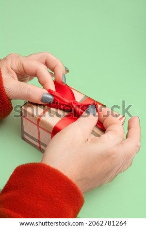 Woman's hands opens a little gift box with a red bow. Light green background. Christmas presents. View from above.