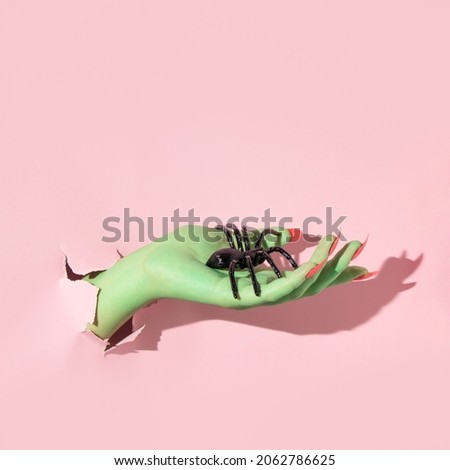 Halloween creative layout with green witch hand with bright pink nails holding spiders against pastel pink background. Aesthetic holiday season  idea. Minimal Halloween concept.