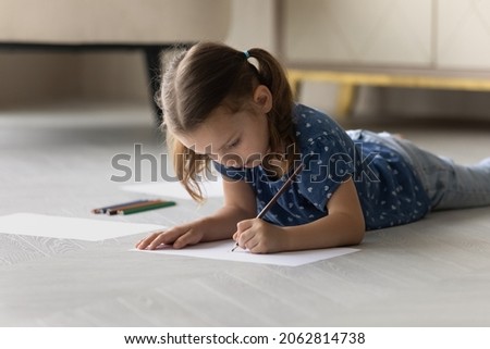 Concentrated happy cute small preschool 6s kid girl lying on warm wooden heated floor, drawing pictures with pencil on paper sheet, enjoying creative hobby pastime activity alone in living room.