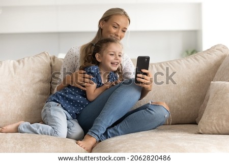 Happy bonding young mother and small cute kid daughter using cellphone, recording funny video for social networks, watching photo content, playing games together at home, online entertainment concept.