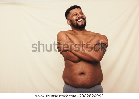 Man with pot belly embracing his natural body. Happy young man laughing cheerfully while standing shirtless against a studio background. Body positive man wearing underwear in a studio. Royalty-Free Stock Photo #2062826963