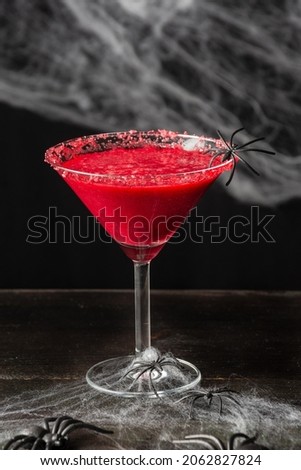 Top view of glass with red halloween cocktail with spider, on wooden table, black background with spider web, vertical