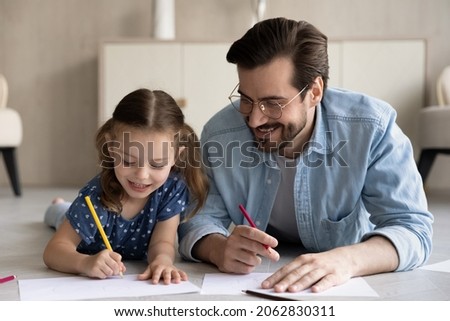 Joyful two different generations family lying on heated wooden floor, involved in drawing pictures on paper sheet, happy small 7s child daughter enjoying creative activity with caring daddy at home.