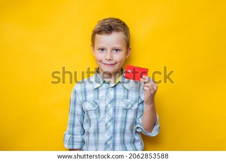Satisfied boy holding a credit bank card isolated on yellow color wall background, child studio portrait
