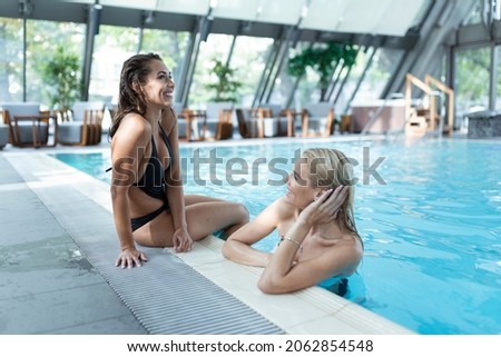 Two girlfriends in swimwear sitting on the poolside, enjoying summer vacation on the swimming pool intdoors