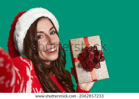 Close up shot of a smiling brunette holding golden gift box with bow knot, lady is ready to celebrate, wears red ugly sweater with deer and santa hat on green background. Christmas concept