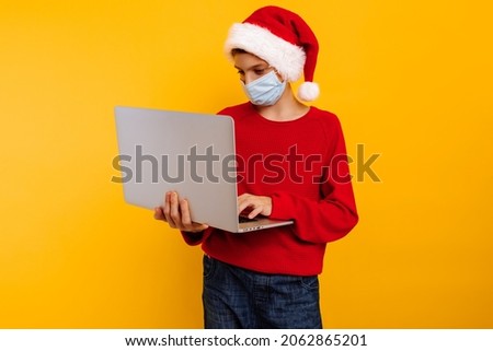 boy in a protective medical mask with a laptop in his hands and a santa claus hat, on a yellow background. communicates by zoom, online with the teacher. distance online learning. christmas concept
