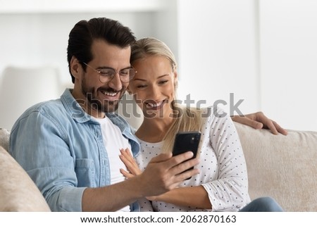 Happy bonding loving married family couple using cellphone, resting on cozy sofa at home. Affectionate young man in eyeglasses cuddling smiling wife, web surfing information or playing games.