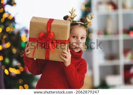 sweet, cheerful, mischievous little girl with a box in her hands, tied with a red ribbon. child is smiling and holding a gift in his hands. Christmas tree in the light of garlands in the background.