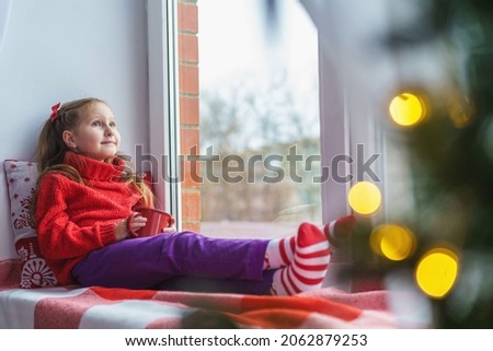 A cute little girl is sitting by the window with a cup of hot drink. Decorated Christmas tree. a child in warm clothes with a blanket and a pillow by the window. Merry Christmas and happy holidays.