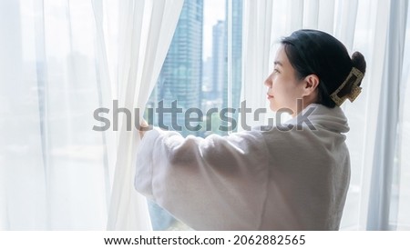 Confident young woman wear nightgown stand open curtain lace looking outside enjoy morning view. Lady wearing shower robe opens white window curtains and looks at the skyscrapers in the big city. Royalty-Free Stock Photo #2062882565