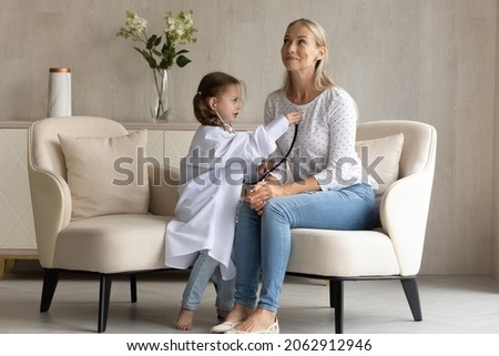 Concentrated happy adorable small child daughter in medical uniform listening heartbeat of beautiful cheerful young mother, playing interesting doctor patient game on weekend in modern living room. Royalty-Free Stock Photo #2062912946