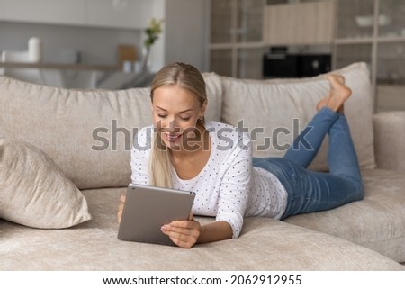 Full length carefree millennial generation beautiful blonde woman using digital touchpad gadget, resting on cozy sofa in modern living room. Happy young lady web surfing information or shopping.