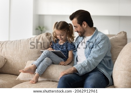 Caring smiling young father in eyeglasses watching happy little preschool kid daughter playing games on digital tablet, resting together on comfortable sofa, parental control, tech addiction concept.