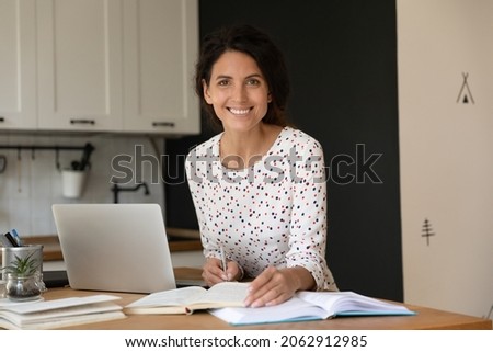 Happy female adult student with laptop and notebooks studying online from home, writing notes, preparing for exam, looking at camera, smiling. Distance learning, education concept. Head shot portrait