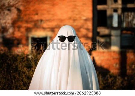 A man in a ghost costume made from a sheet and sunglasses stands near an abandoned building. Ghost Challenge 2021. Spooky season. Celebrating halloween.