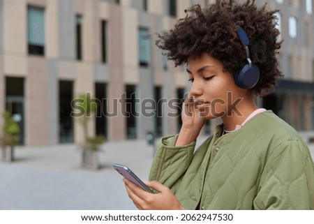 Profile shot of curly haired woman reads news via mobile phone wears wireless headphones listens audio track from playlist dressed in jacket stands against modern building checks social networks