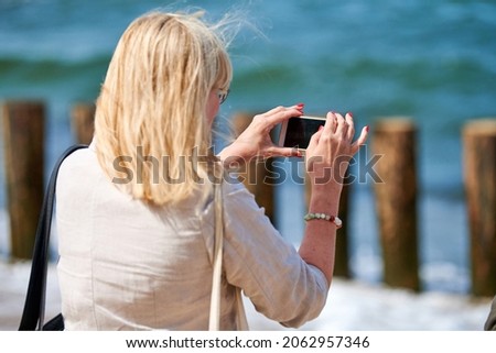 Blonde woman taking photos on phone, back view. Shooting video with cell phone camera, using smartphone to take pictures outdoor. Tourist photographing landscape by sea. Sightseeing concept