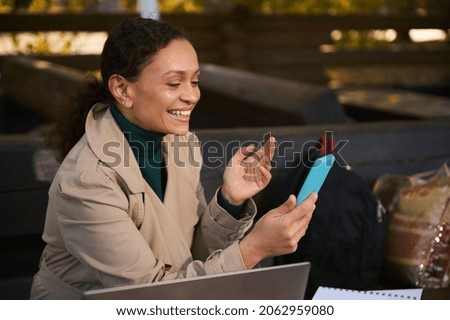 Cheerful attractive woman smiles while making a video call via a mobile app on her smartphone. Young confident business woman working outdoors away from urban civilization on the oak grove autumn cafe