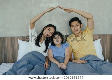 Happy Asian family sitting on bed in bedroom together and making the home sign. Mom and dad making roof figure with hands arms over heads