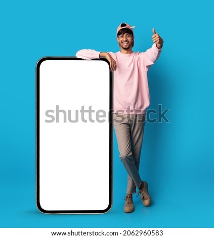 Positive stylish indian guy posing with big smartphone with empty screen, showing thumb up and smiling, recommending newest mobile app, blue studio background, mockup, full length shot Royalty-Free Stock Photo #2062960583