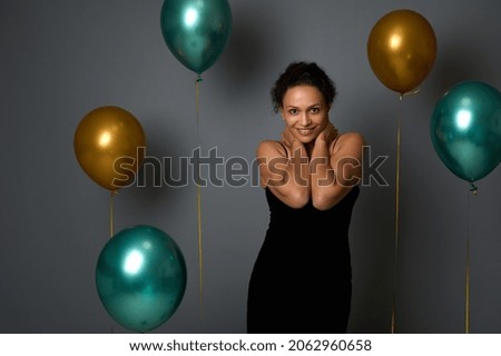 Portrait of cheerful pretty woman in evening black dress smiling with beautiful toothy smile posing against air balloons on gray wall background with copy space for Christmas, New Year, anniversary ad