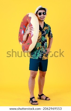 Stylish young Asian man with sunglasses holds colorful swim ring. Half body studio portrait on yellow background. Summer vacations start concept