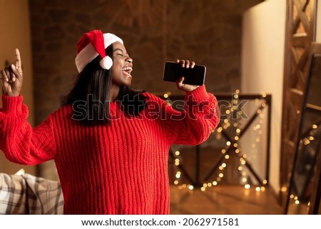 Christmas holiday music. Happy black lady in Santa hat singing, using smartphone as mic at home, mockup for mobile app. Cheerful young woman enjoying karaoke song, having fun on Xmas Royalty-Free Stock Photo #2062971581