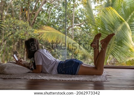 young woman lying on wooden floor, relaxing using smartphone with forest view.	