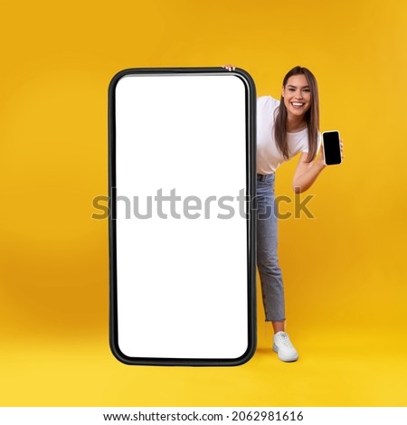 Full Body Length Of Cheerful Woman Peeking Out Standing Behind Big Smartphone With White Blank Screen, Excited Lady Holding Cell Phone Presenting New App Showing Copy Space For Website Design Mock Up Royalty-Free Stock Photo #2062981616