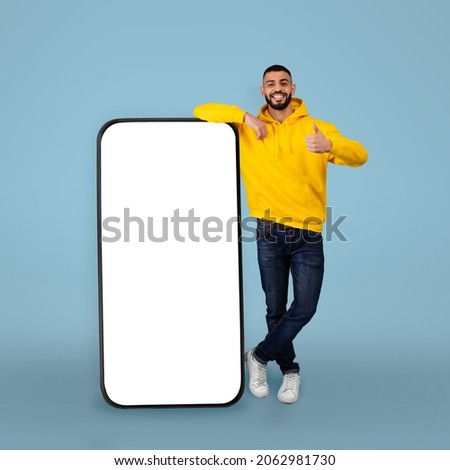 Mobile offer. Happy arab man leaning on big smartphone with blank screen and gesturing thumb up, handsome guy recommending new app or website over blue background, mock up image