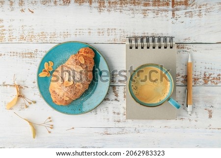 Fresh baked almond nuts breakfast croissant on vintage turquoise plate and hot coffee cup over scrapbook with bamboo pen on rustic wooden background. Top view pastry good morning card with copy space
