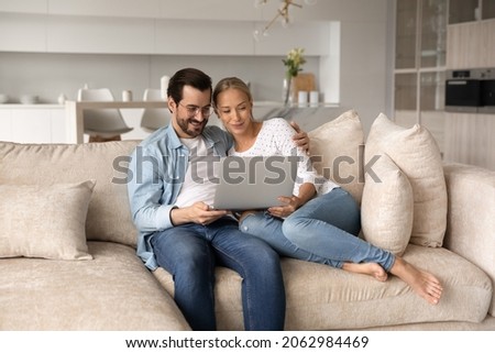 Happy loving young family couple looking at laptop screen, sitting on cozy sofa at home, enjoying watching comedian movie, web surfing information or spending time online in social network on weekend.