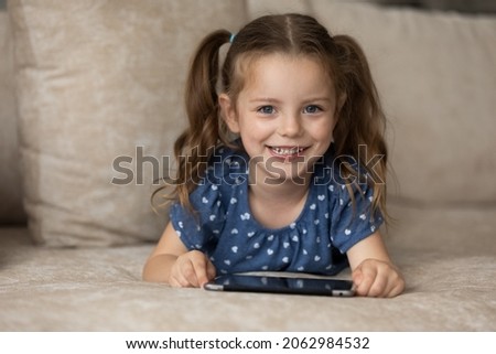 Portrait of adorable little preschool kid girl lying on comfortable sofa with modern gadget cellphone, playing mobile games, entertaining online, secure internet content for children, tech addiction.